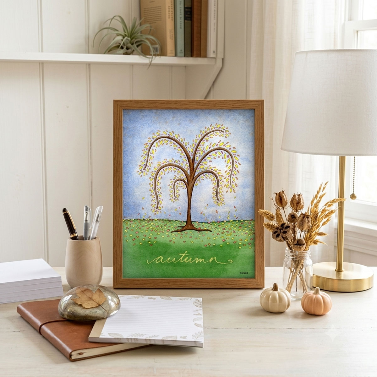 Printable autumn tree wall art featuring a watercolor tree with yellow and orange fall leaves dropping onto a green hill framed and sitting on a desk