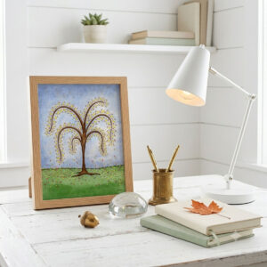 Printable autumn tree wall art with featuring a watercolor tree with yellow and orange fall leaves dropping onto a green hill framed and sitting on a desk