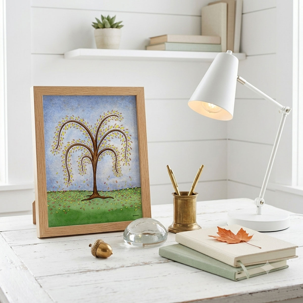 Printable autumn tree wall art with featuring a watercolor tree with yellow and orange fall leaves dropping onto a green hill framed and sitting on a desk
