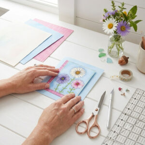 A mock-up photo of a room showing a framed print of a Trio of Daisies Floral Wall Art featuring white, purple, and pink flowers on a blue background with a pink gingham border.