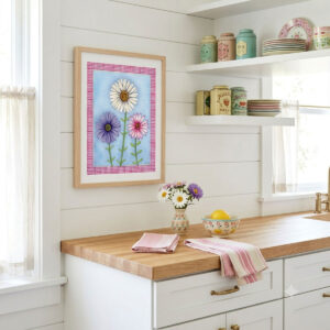 A mock-up photo of a room showing a framed print of a Trio of Daisies Floral Wall Art featuring white, purple, and pink flowers on a blue background with a pink gingham border.