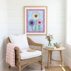 A mock-up photo of a room showing a framed print of a Trio of Daisies Floral Wall Art featuring white, purple, and pink flowers on a blue background with a pink gingham border.